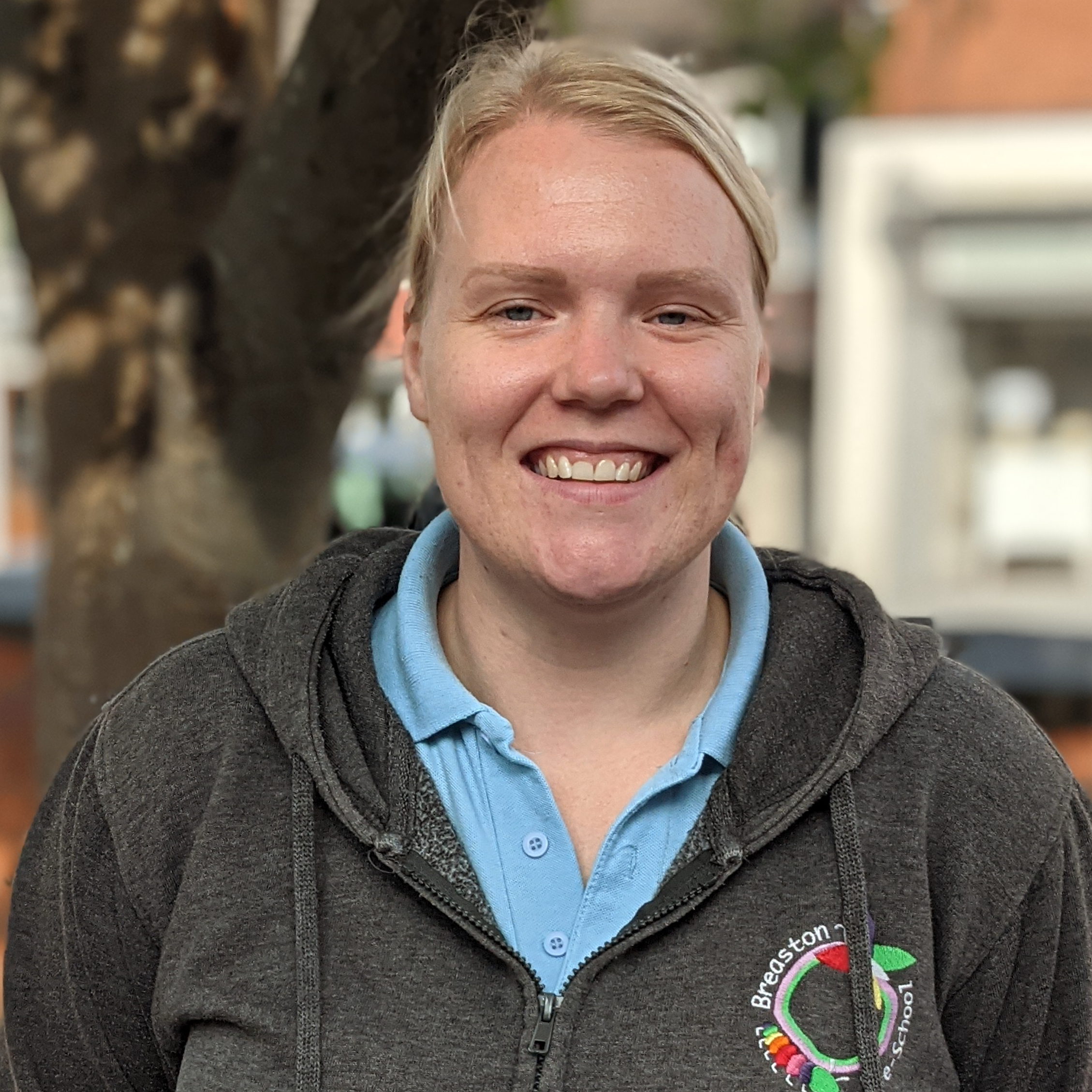 Hayley, a white woman with blonde hair tied back, is smiling at the camera. She is wearing a grey hoodie with the Pre-School logo visible over a blue tshirt.